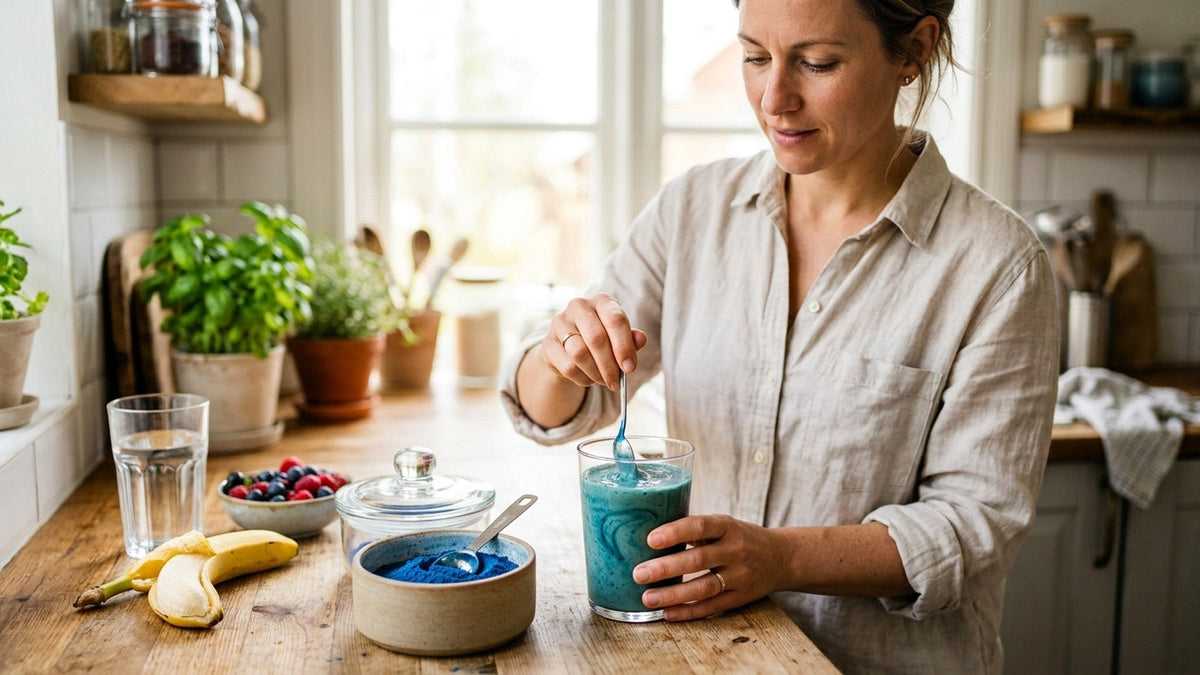 An adult stirs blue spirulina powder into a fruit smoothie at a bright kitchen counter.