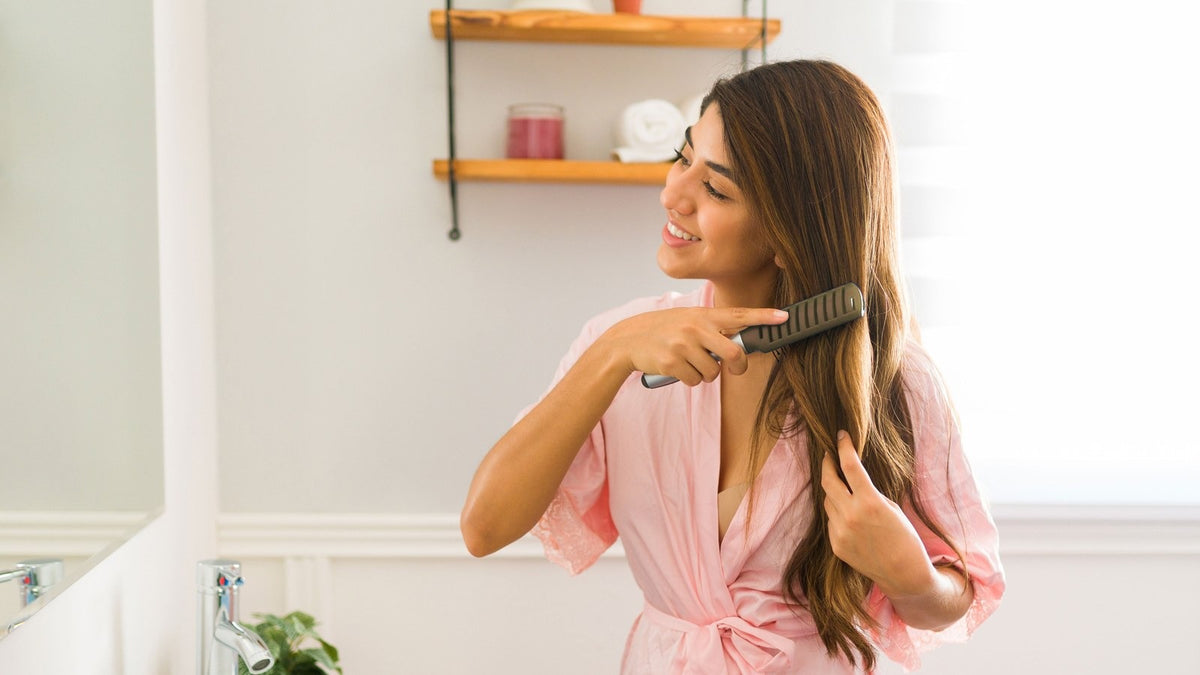 Young woman with long brown hair in a pink robe brushing her hair in a bright bathroom at home