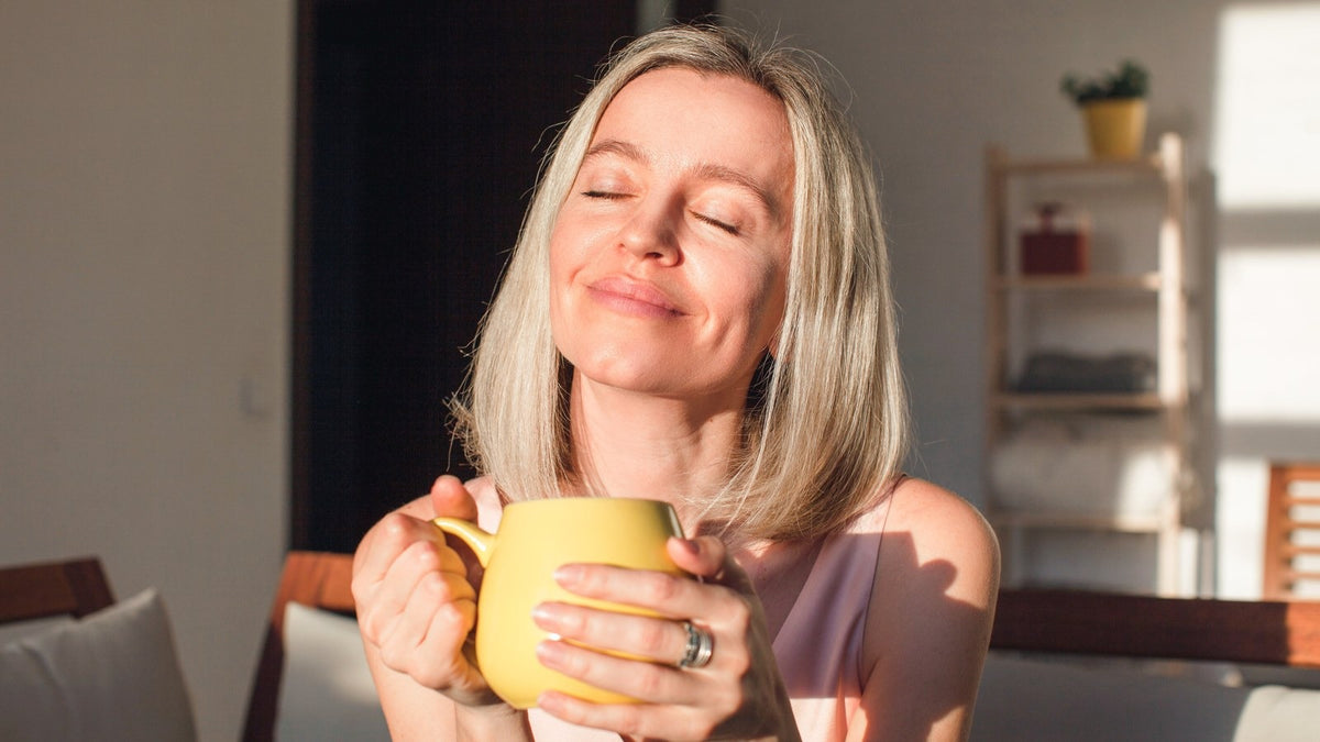 Woman with shoulder-length blonde hair holds a yellow mug at home, eyes closed in morning sun, enjoying her magnesium routine